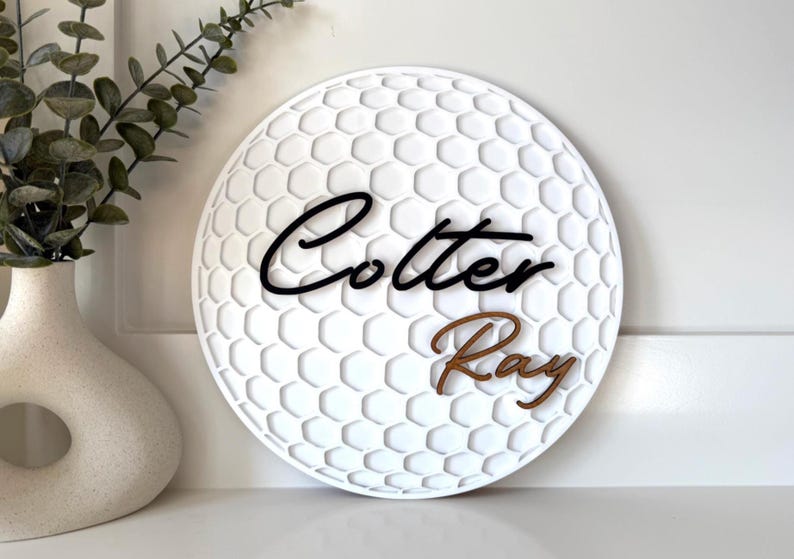 May include: White, round wall decor with a golf ball pattern. The name "Cotter Ray" is displayed in black and brown script. Decorative item for a home.