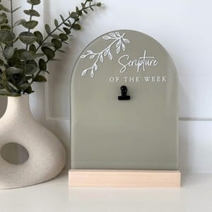May include: A sage green arched acrylic sign with the text "Scripture of the Week" and a small black clip. The sign is mounted on a light wood base. A white vase with a unique shape and green plant are to the left.