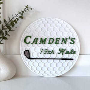 May include: A circular white wall sign textured like a golf ball surface. It features the dark green text "CAMDEN'S 19th Hole" and a black golf club graphic positioned horizontally across the lower section of the sign.