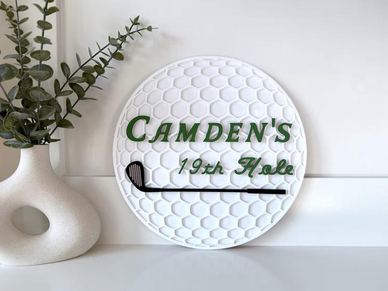 May include: A circular white wall sign textured like a golf ball surface. It features the dark green text "CAMDEN'S 19th Hole" and a black golf club graphic positioned horizontally across the lower section of the sign.