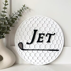 May include: A circular white wall sign featuring a textured honeycomb pattern resembling a golf ball. The sign displays the black text "Jet" in a serif font positioned above a black silhouette of a golf club, creating a decorative sports-themed piece.