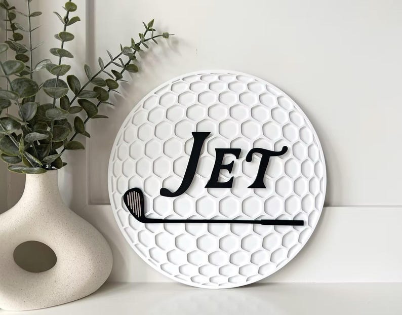 May include: A circular white wall sign featuring a textured honeycomb pattern resembling a golf ball. The sign displays the black text "Jet" in a serif font positioned above a black silhouette of a golf club, creating a decorative sports-themed piece.