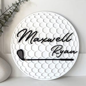 May include: A circular white wall sign featuring a textured honeycomb pattern resembling a golf ball. The sign displays the black script text Maxwell Ryan and a black golf club graphic positioned horizontally across the lower section.