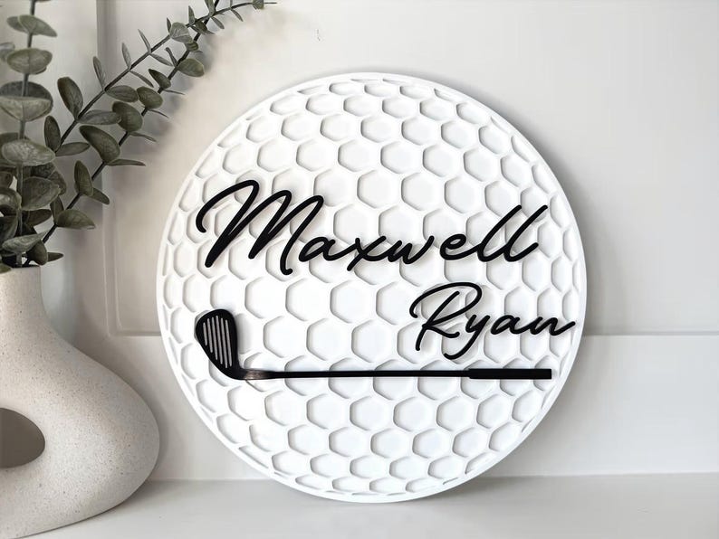 May include: A circular white wall sign featuring a textured honeycomb pattern resembling a golf ball. The sign displays the black script text Maxwell Ryan and a black golf club graphic positioned horizontally across the lower section.