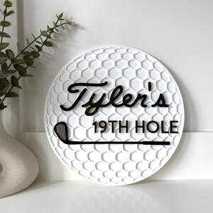 May include: A circular white wall sign with a textured golf ball dimple pattern. The sign features black raised lettering that reads Tyler's 19TH HOLE and a black graphic of a golf club positioned horizontally across the bottom section.