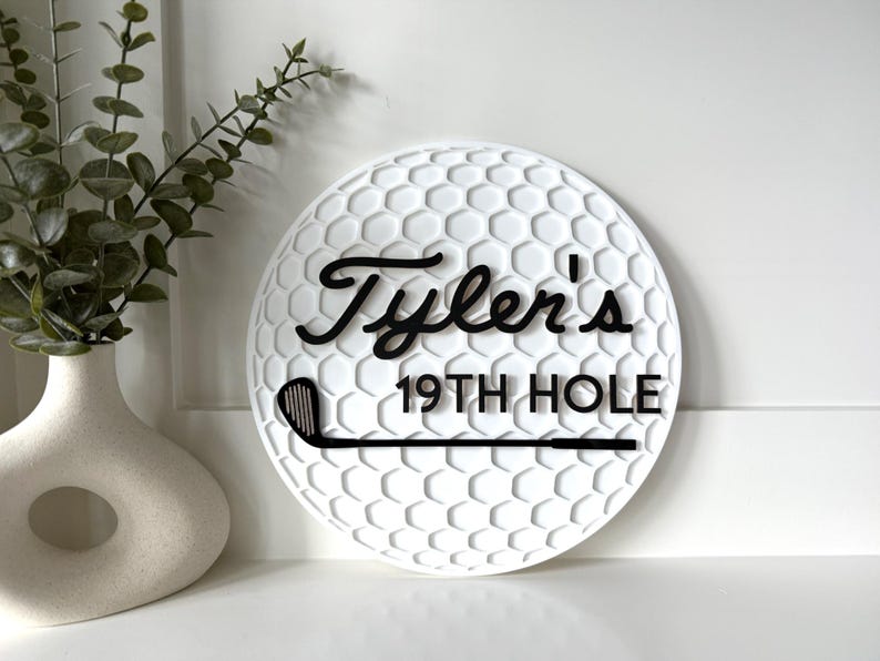 May include: A circular white wall sign with a textured golf ball dimple pattern. The sign features black raised lettering that reads Tyler's 19TH HOLE and a black graphic of a golf club positioned horizontally across the bottom section.