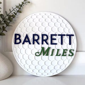 May include: White golf ball-shaped sign with a textured surface and the names "BARRETT" in navy blue and "MILES" in green. The sign is displayed near a white vase with greenery.