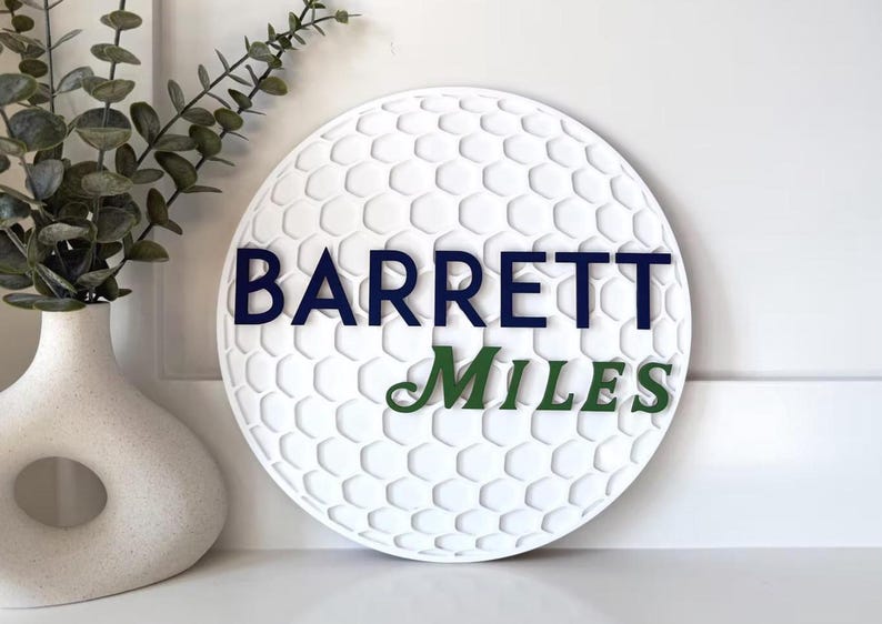 May include: White golf ball-shaped sign with a textured surface and the names "BARRETT" in navy blue and "MILES" in green. The sign is displayed near a white vase with greenery.