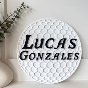 May include: A white, round sign with a golf ball pattern and the name "Lucas Gonzales" in black lettering. The sign is displayed near a decorative vase with greenery.