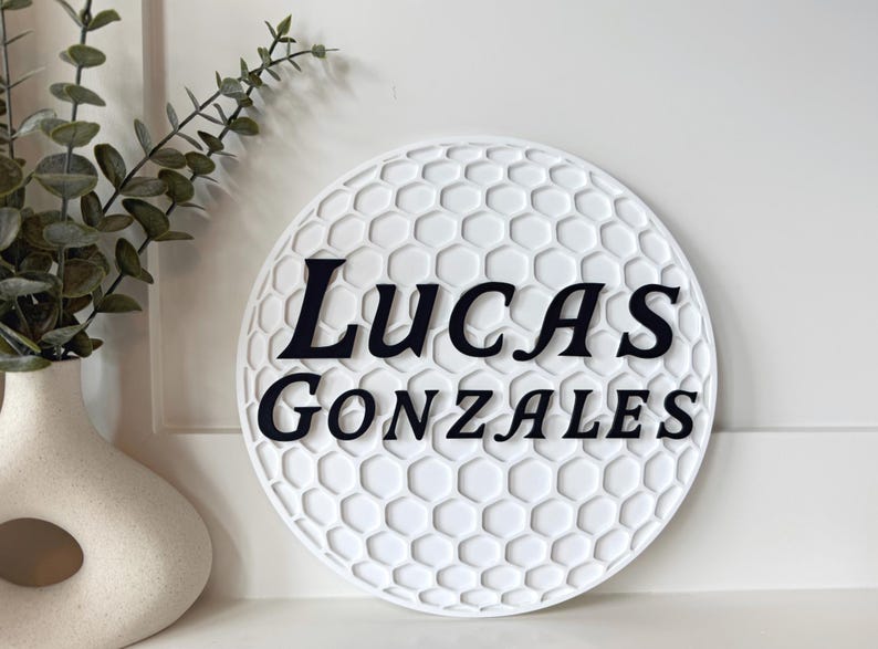 May include: A white, round sign with a golf ball pattern and the name "Lucas Gonzales" in black lettering. The sign is displayed near a decorative vase with greenery.