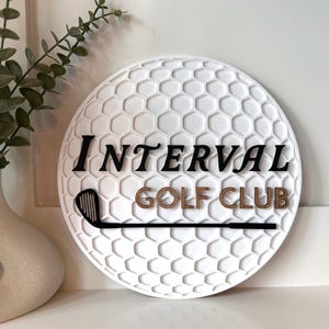 May include: A circular white wall sign with a textured honeycomb pattern resembling a golf ball. The sign features the black text "INTERVAL" above the brown text "GOLF CLUB" and a black silhouette of a golf club iron positioned horizontally.