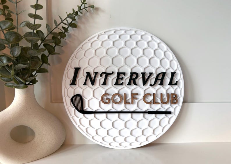 May include: A circular white wall sign with a textured honeycomb pattern resembling a golf ball. The sign features the black text "INTERVAL" above the brown text "GOLF CLUB" and a black silhouette of a golf club iron positioned horizontally.