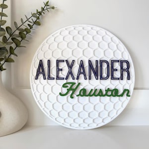May include: A white, round sign with a golf ball pattern. The name "ALEXANDER" is in navy blue letters, and "Houston" is in green script. A decorative vase with greenery is in the background.