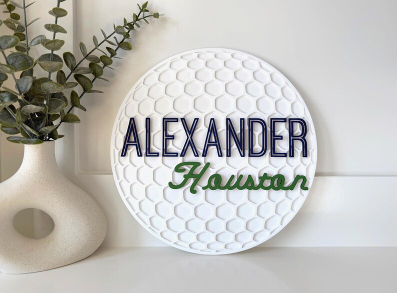 May include: A white, round sign with a golf ball pattern. The name "ALEXANDER" is in navy blue letters, and "Houston" is in green script. A decorative vase with greenery is in the background.