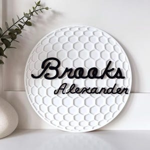 May include: White, round decorative sign with a golf ball pattern. The name "Brooks Alexander" is written in black cursive script across the sign. A white vase with greenery is in the background.