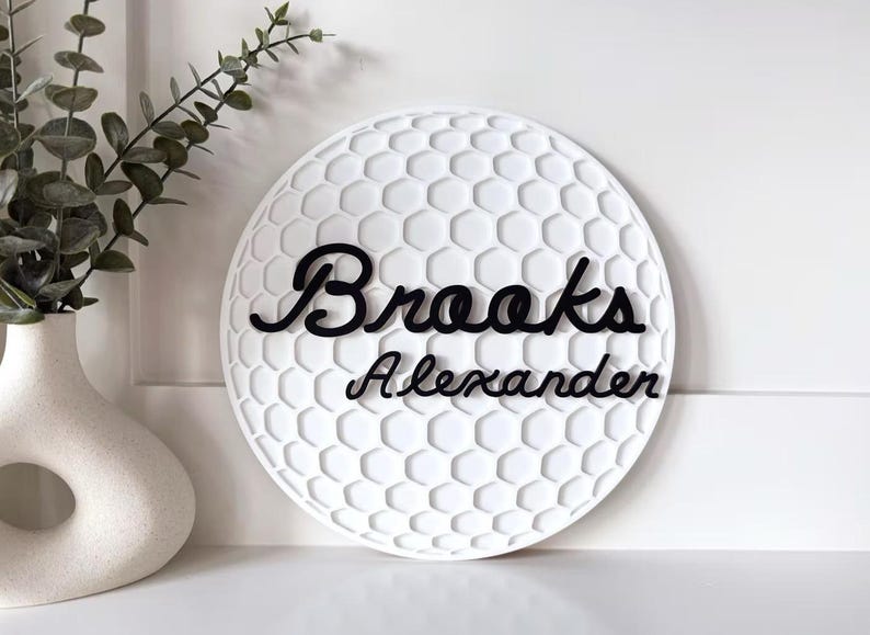 May include: White, round decorative sign with a golf ball pattern. The name "Brooks Alexander" is written in black cursive script across the sign. A white vase with greenery is in the background.