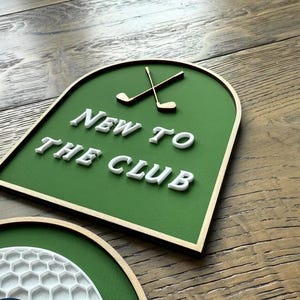 May include: An arched sign with a green background and a thin gold border. The sign features two crossed gold golf clubs at the top and the white raised text NEW TO THE CLUB centred below. A partial circular sign with a white honeycomb pattern is visible.