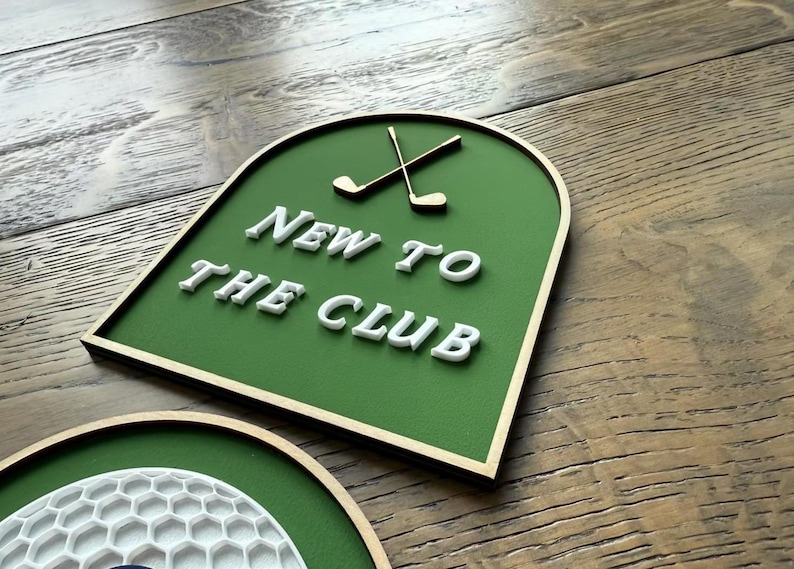 May include: An arched sign with a green background and a thin gold border. The sign features two crossed gold golf clubs at the top and the white raised text NEW TO THE CLUB centred below. A partial circular sign with a white honeycomb pattern is visible.