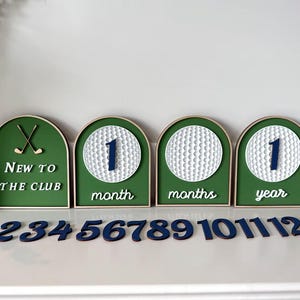 May include: A set of four arched green signs with white golf ball designs and dark blue numbers. The signs feature the text New to the club, month, months, and year. A separate set of dark blue wooden numbers from 2 to 12 is included.