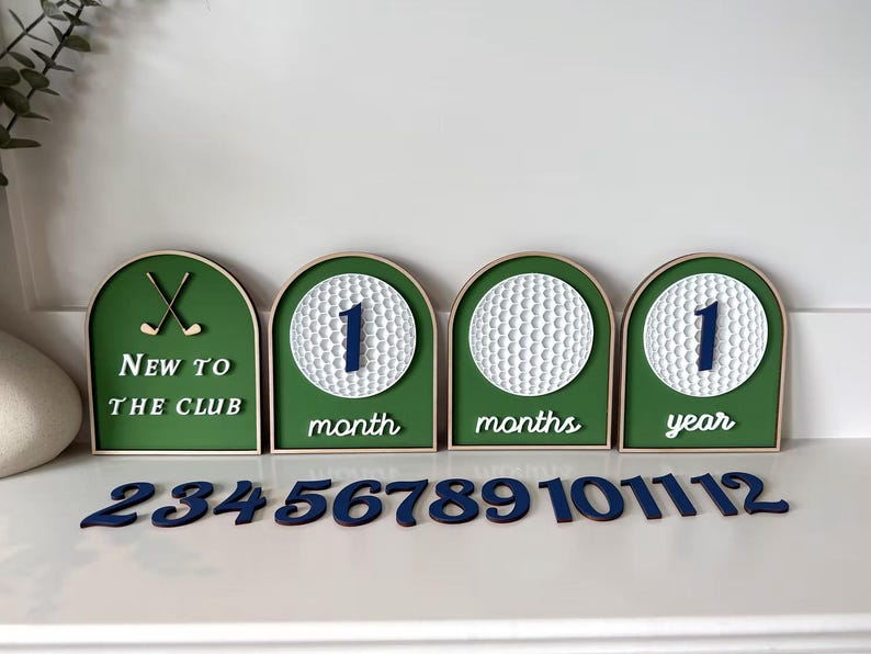 May include: A set of four arched green signs with white golf ball designs and dark blue numbers. The signs feature the text New to the club, month, months, and year. A separate set of dark blue wooden numbers from 2 to 12 is included.