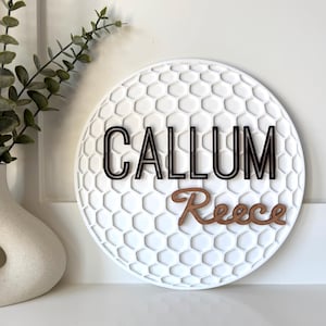 May include: A white, round wall decor piece with a golf ball texture. The name "CALLUM" is in black, and "Reece" is in brown. A white vase with greenery is to the left.