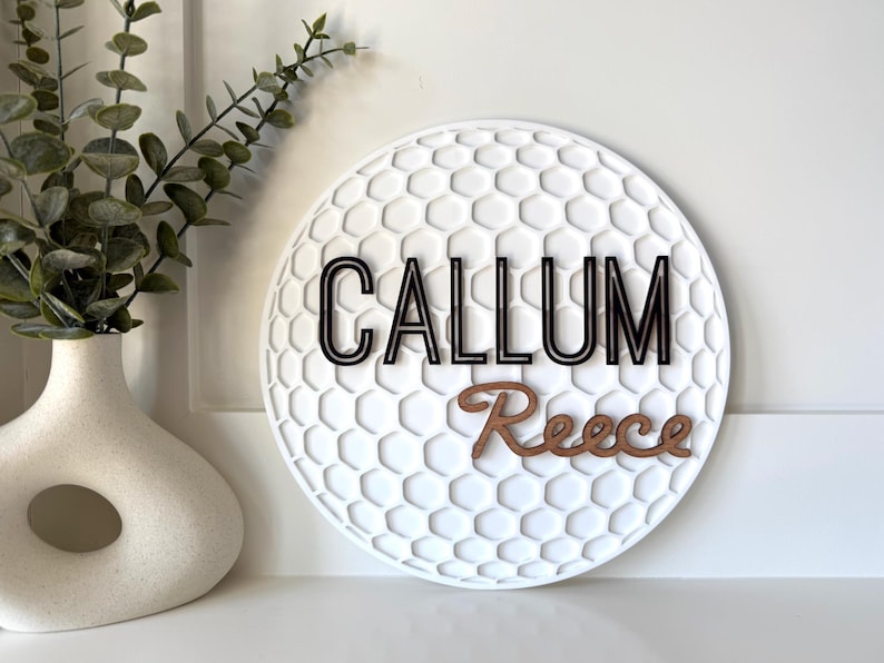 May include: A white, round wall decor piece with a golf ball texture. The name "CALLUM" is in black, and "Reece" is in brown. A white vase with greenery is to the left.