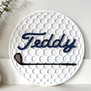 May include: A circular white wall sign featuring a textured honeycomb pattern resembling a golf ball. The name Teddy is written in dark blue cursive script across the center, with a black golf club icon positioned horizontally below the name.