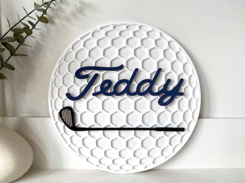 May include: A circular white wall sign featuring a textured honeycomb pattern resembling a golf ball. The name Teddy is written in dark blue cursive script across the center, with a black golf club icon positioned horizontally below the name.
