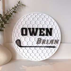 May include: A circular white wall sign featuring a textured honeycomb pattern resembling a golf ball. The sign displays the name OWEN in bold black letters above the name BRIAN in grey, accompanied by a black silhouette of a golf club.