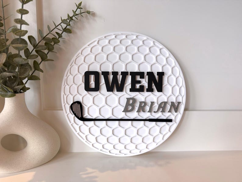 May include: A circular white wall sign featuring a textured honeycomb pattern resembling a golf ball. The sign displays the name OWEN in bold black letters above the name BRIAN in grey, accompanied by a black silhouette of a golf club.