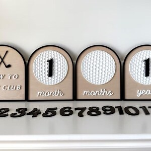 May include: Four arched wooden milestone signs with black borders. One sign features crossed golf clubs and the text "NEW TO THE CLUB". Three signs feature white golf ball textures with interchangeable black numbers and white text "month", "months", or "year".