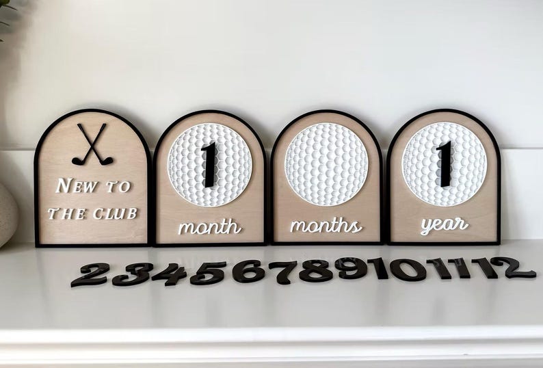 May include: Four arched wooden milestone signs with black borders. One sign features crossed golf clubs and the text "NEW TO THE CLUB". Three signs feature white golf ball textures with interchangeable black numbers and white text "month", "months", or "year".