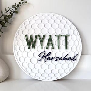 May include: A white, round sign with a golf ball texture. The name "WYATT" is in green letters above the name "Herschel" in a dark blue script. A decorative vase with greenery is in the background.