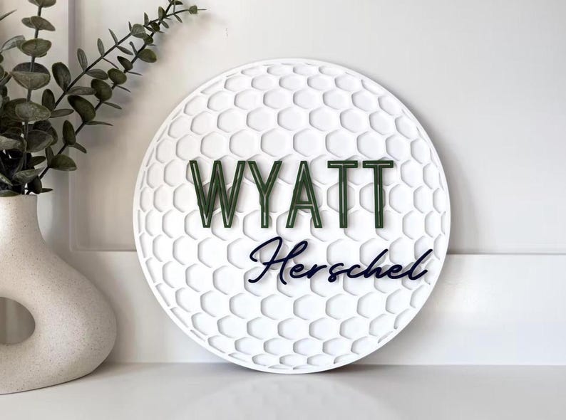 May include: A white, round sign with a golf ball texture. The name "WYATT" is in green letters above the name "Herschel" in a dark blue script. A decorative vase with greenery is in the background.