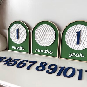 May include: Three arched wooden signs with green backgrounds and white golf ball textures. The signs display the text 1 month, months, and 1 year. A set of separate dark blue wooden numbers from 2 to 11 is arranged in front of the signs.