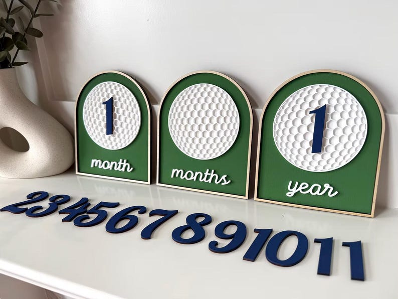 May include: Three arched wooden signs with green backgrounds and white golf ball textures. The signs display the text 1 month, months, and 1 year. A set of separate dark blue wooden numbers from 2 to 11 is arranged in front of the signs.