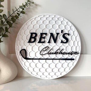 May include: A circular white sign with a textured golf ball dimple pattern. The sign features the black text "BEN'S" in a bold serif font above the black script text "Clubhouse" and a black graphic of a golf club head and shaft.