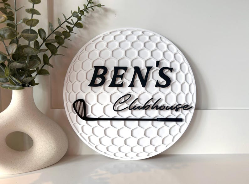 May include: A circular white sign with a textured golf ball dimple pattern. The sign features the black text "BEN'S" in a bold serif font above the black script text "Clubhouse" and a black graphic of a golf club head and shaft.