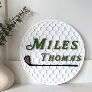 May include: A circular white wall sign with a textured honeycomb pattern resembling a golf ball. The sign features the dark green text "MILES THOMAS" in a serif font and a black golf club silhouette positioned horizontally below the name.