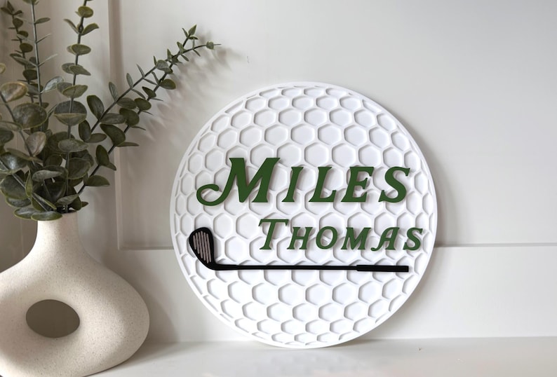 May include: A circular white wall sign with a textured honeycomb pattern resembling a golf ball. The sign features the dark green text "MILES THOMAS" in a serif font and a black golf club silhouette positioned horizontally below the name.