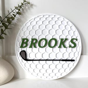 May include: A circular white wall sign featuring a textured honeycomb pattern resembling a golf ball. The sign displays the name BROOKS in dark green capital letters and includes a black silhouette of a golf club positioned horizontally below.