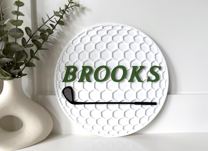 May include: A circular white wall sign featuring a textured honeycomb pattern resembling a golf ball. The sign displays the name BROOKS in dark green capital letters and includes a black silhouette of a golf club positioned horizontally below.