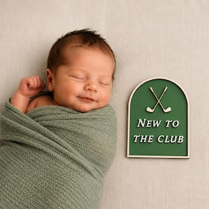May include: A sleeping newborn wrapped in a sage green knit swaddle next to an arched wooden sign with a dark green background, two crossed golf clubs, and the white text NEW TO THE CLUB, featuring a light wood frame and base.
