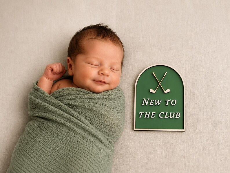 May include: A sleeping newborn wrapped in a sage green knit swaddle next to an arched wooden sign with a dark green background, two crossed golf clubs, and the white text NEW TO THE CLUB, featuring a light wood frame and base.