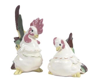 May include: A pair of ceramic rooster and hen figurines. The rooster stands upright with a pink comb and wattle, while the hen is in a seated position. Both are primarily white with accents of green, brown, and yellow.