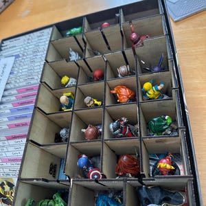 May include: A wooden box with compartments for storing miniature figures. The box is filled with various figures, including superheroes, villains, and other characters. The box is labeled with the text "Room for 15 Locs".