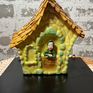Peut inclure: Un pot de fleurs en céramique jaune en forme de chalet avec un toit vert et une petite figurine verte qui regarde par la porte. Le pot a une porte brune et une fenêtre avec un motif en losange.