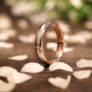 Puede incluir: Una banda de boda de oro rosa y blanco con una banda blanca delgada alrededor del centro del anillo.