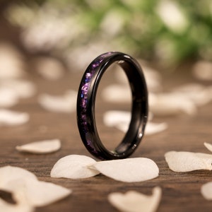 May include: A black tungsten carbide ring with a purple and white inlay. The ring is shown on a wooden surface with white flower petals.