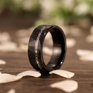 Può includere: Un anello in carburo di tungsteno nero con finitura martellata e una fila di piccoli diamanti incastonati nella fascia.